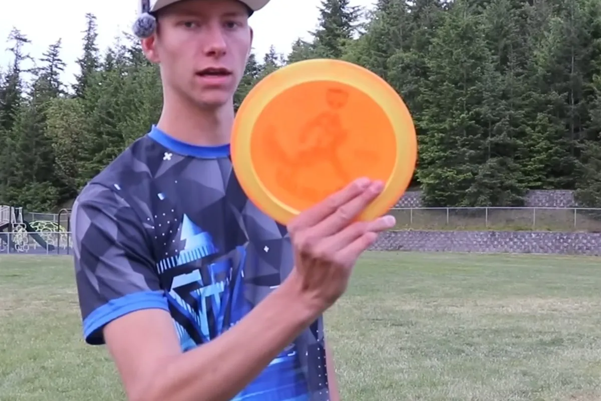 Gannon Buhr holding up an orange disc in his right hand to show his forehand grip, with his fingers pressed against the flight plate