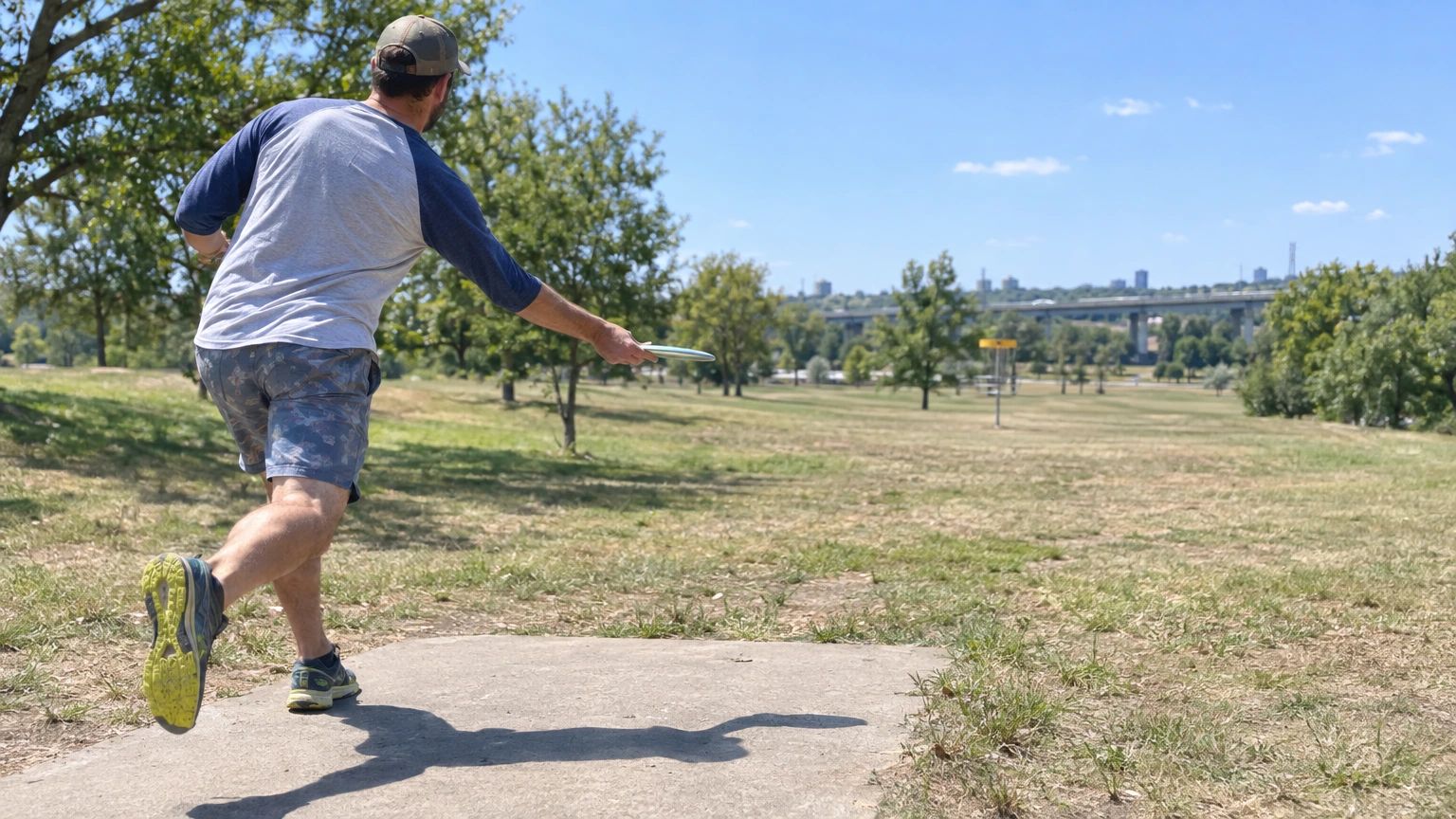A disc golfer seen from behind in the middle of a forehand throw on a concrete tee pad in a sunny park, the disc flat in his hand and a basket far down the fairway.